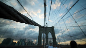 Brooklyn Bridge close-up of suspension cables, people crossing on pedestrian walkway at sunset with Manhattan skyline