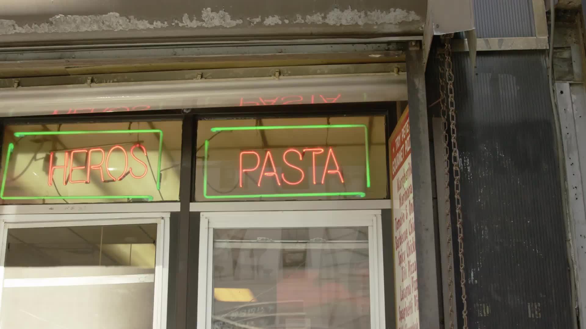 neon heros and pasta sign in pizzeria deli shop with cook in uniform in window