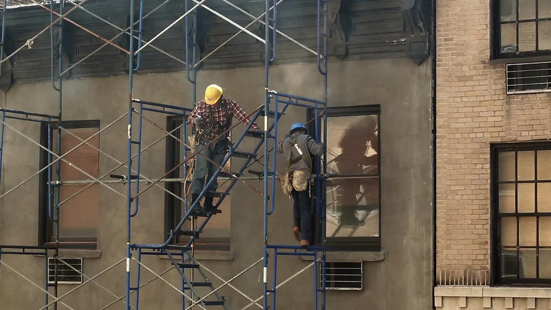 construction workers with yellow hardhats on scaffolding climbing side of building to roof at top