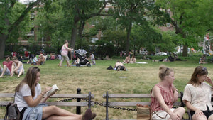 women reading on benches in Washington Square Park in summer with green trees in background and people sunbathing