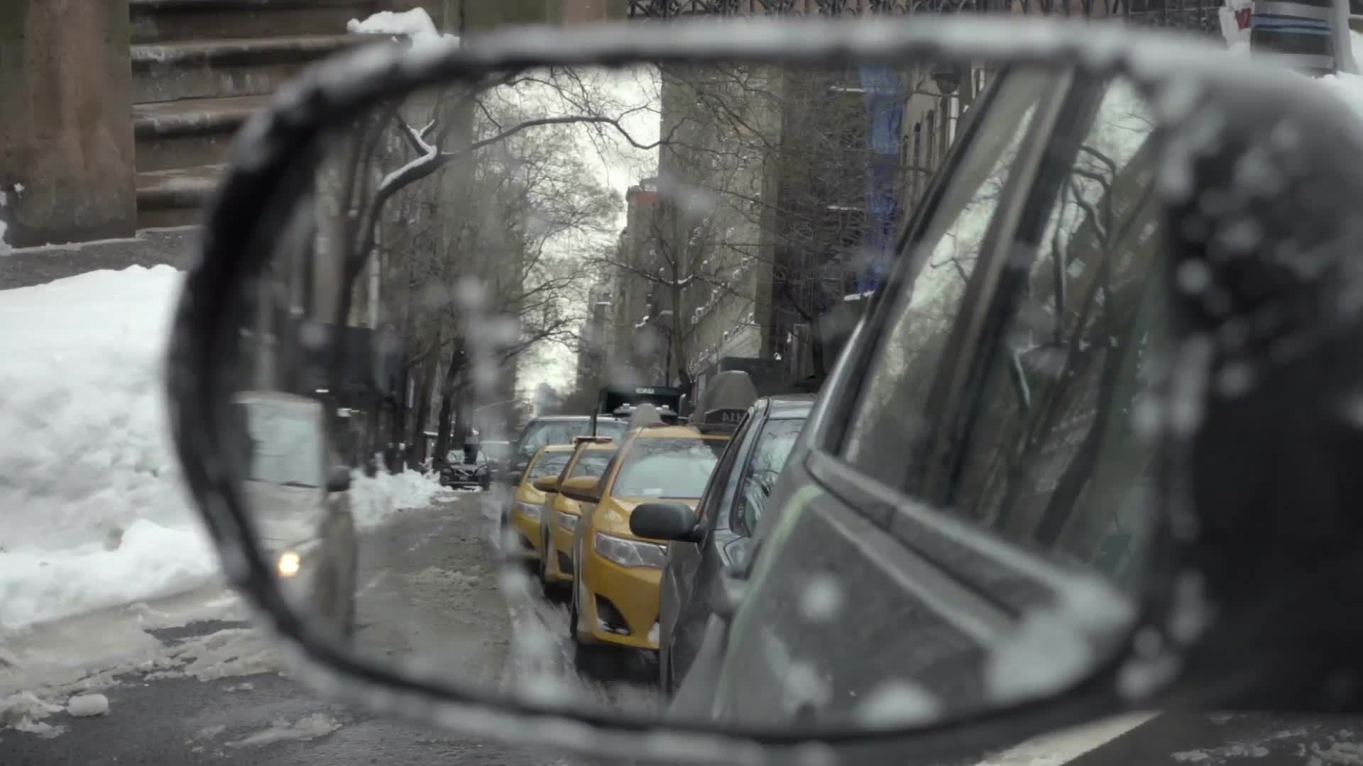 taxi cabs and cars in driver's side mirror while driving in slow motion on winter day with snow on ground in NYC